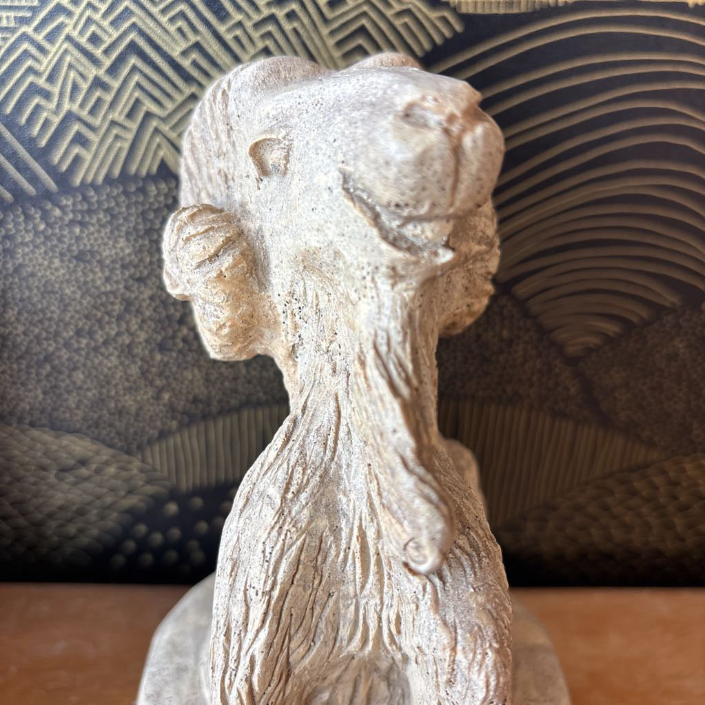 A carved stone figure of a ram on a pedestal. It sits on a wooden surface and a black and gold wallpaper backdrop.