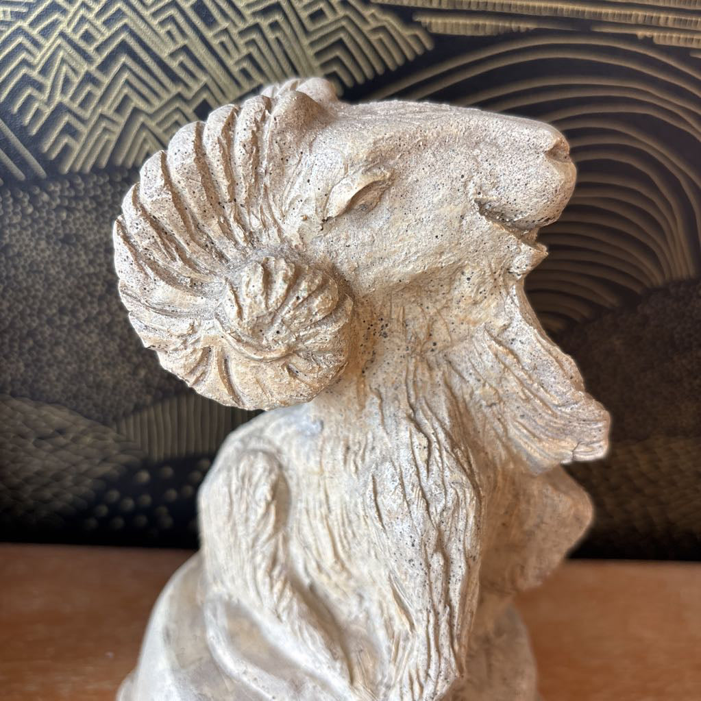 A carved stone figure of a ram on a pedestal. It sits on a wooden surface and a black and gold wallpaper backdrop.