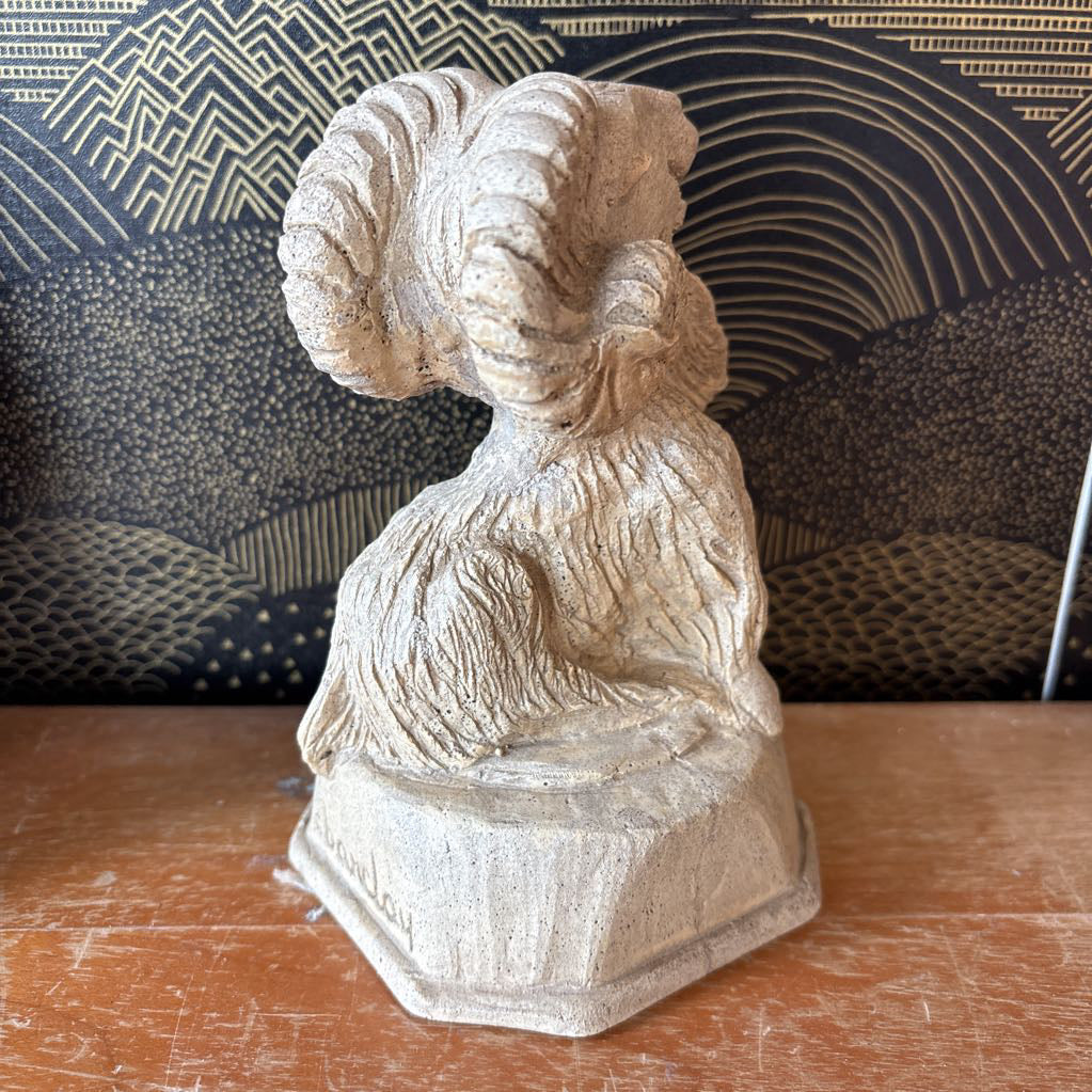 A small beige stone or concrete ram figurine with large curled horns, standing on a hexagonal base on a wooden tabletop. A patterned dark blue and gold cushion is visible in the background.