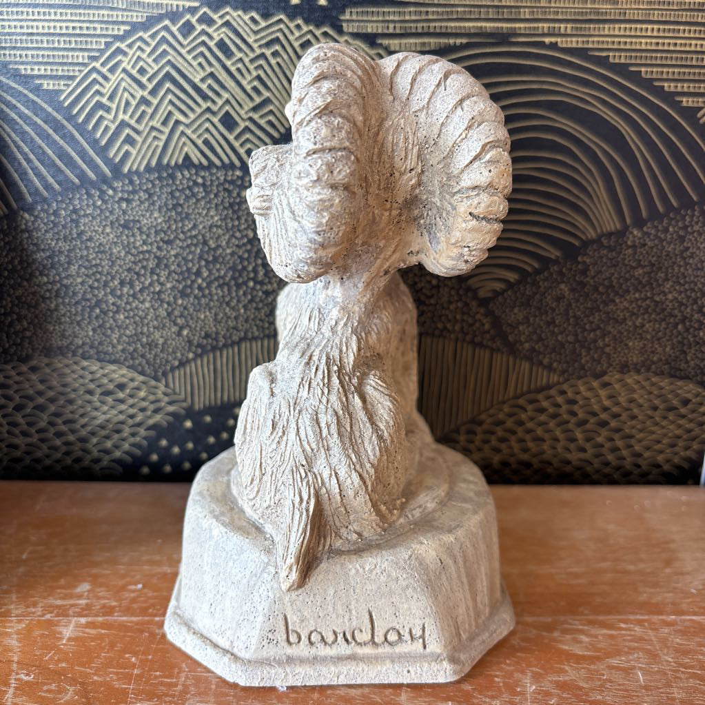 A carved stone figure of a ram on a pedestal. It sits on a wooden surface and a black and gold wallpaper backdrop.