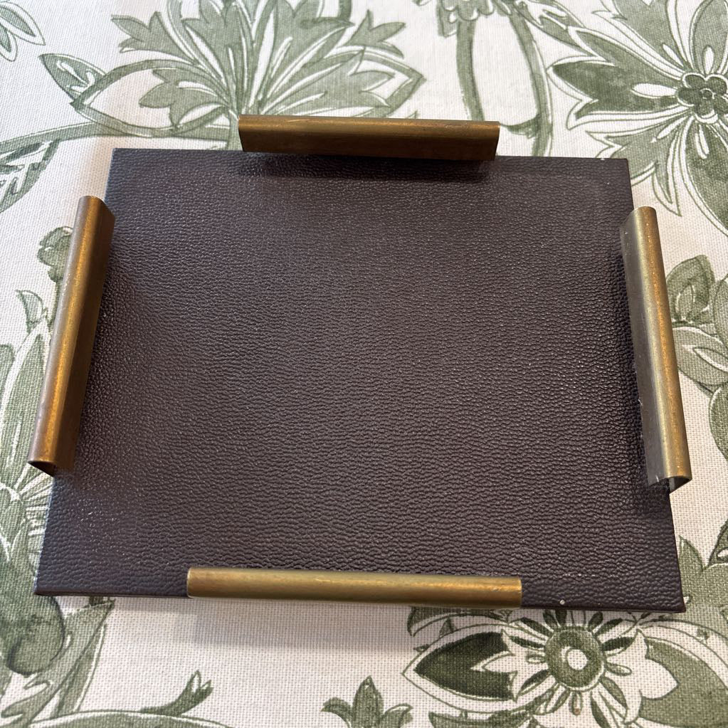 A square dark brown textured tray with four cylindrical handles in a brass-like metallic finish, placed on a green and white floral patterned fabric.