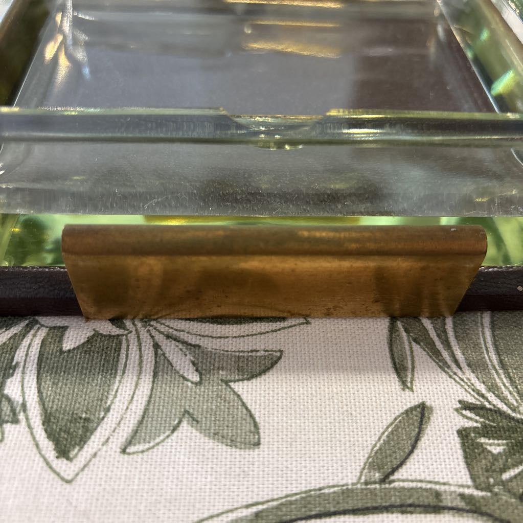 A close-up of a rectangular brass drawer pull handle mounted on a dark drawer front, positioned above a white fabric surface with a green floral pattern.