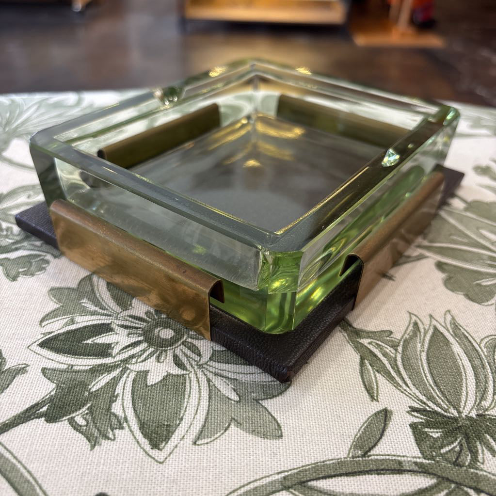 A square glass ashtray with a translucent green-tinted glass body and a dark base, photographed on a floral tablecloth.
