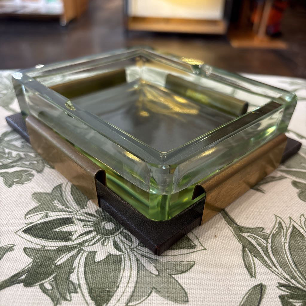 A square glass ashtray with a thick clear glass rim, resting on a brown wooden base with black textured corner supports, set on a green and white floral tablecloth.