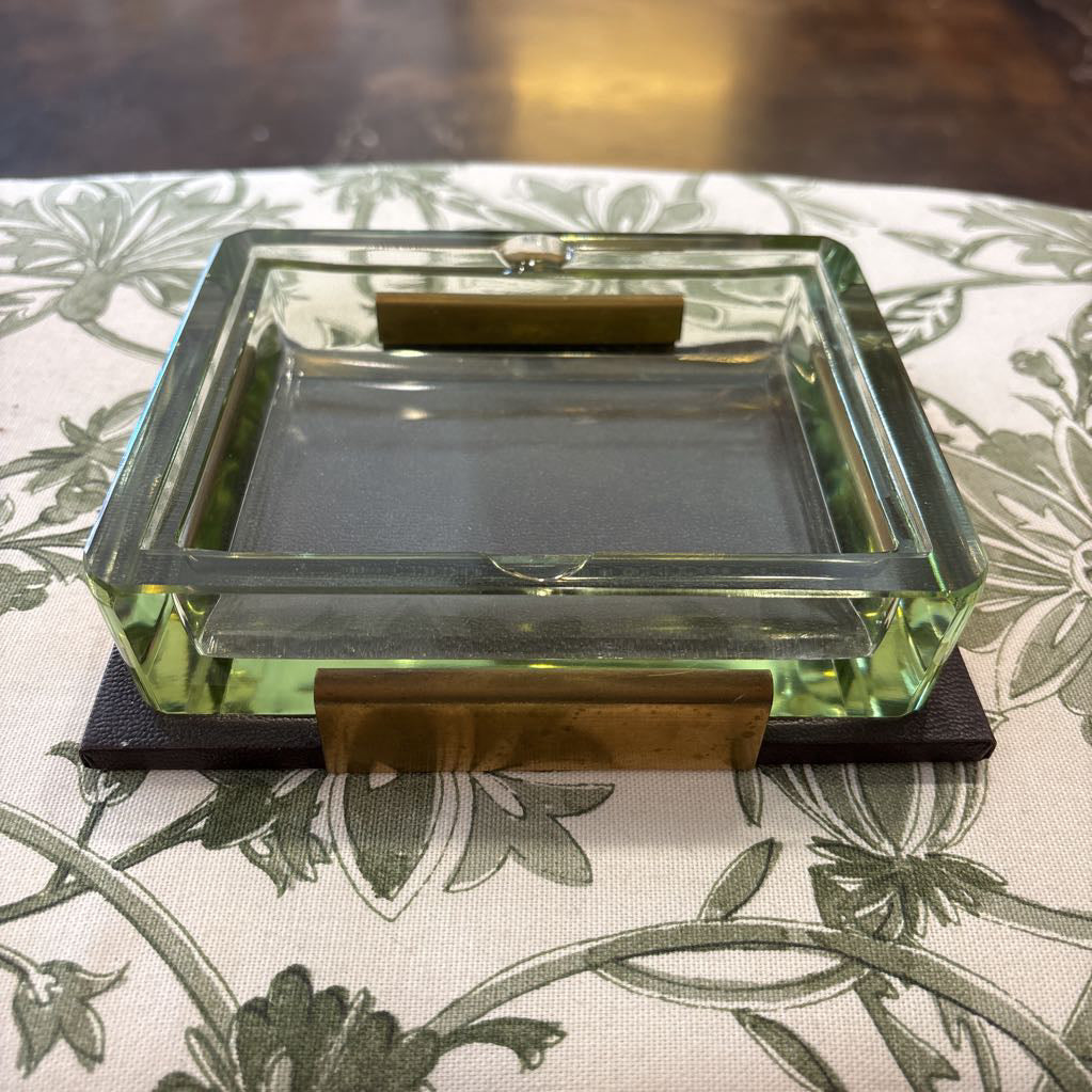 A square clear glass ashtray with a light green tinted base, featuring two brass metal accents on the front, resting on a black coaster over a floral tablecloth.