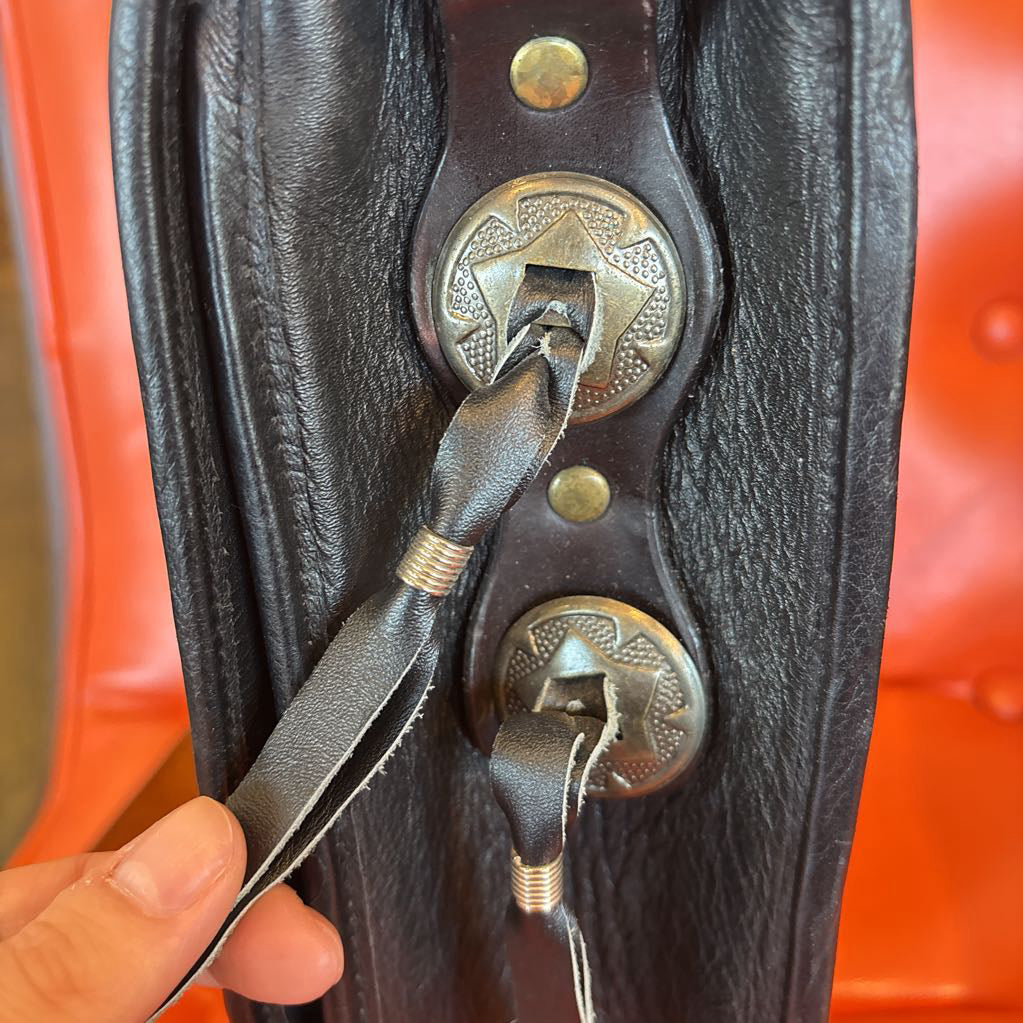 A black leather belt bag with silver hardware and tied leather pieces. This bag sits on an orange cushion in a room setting.