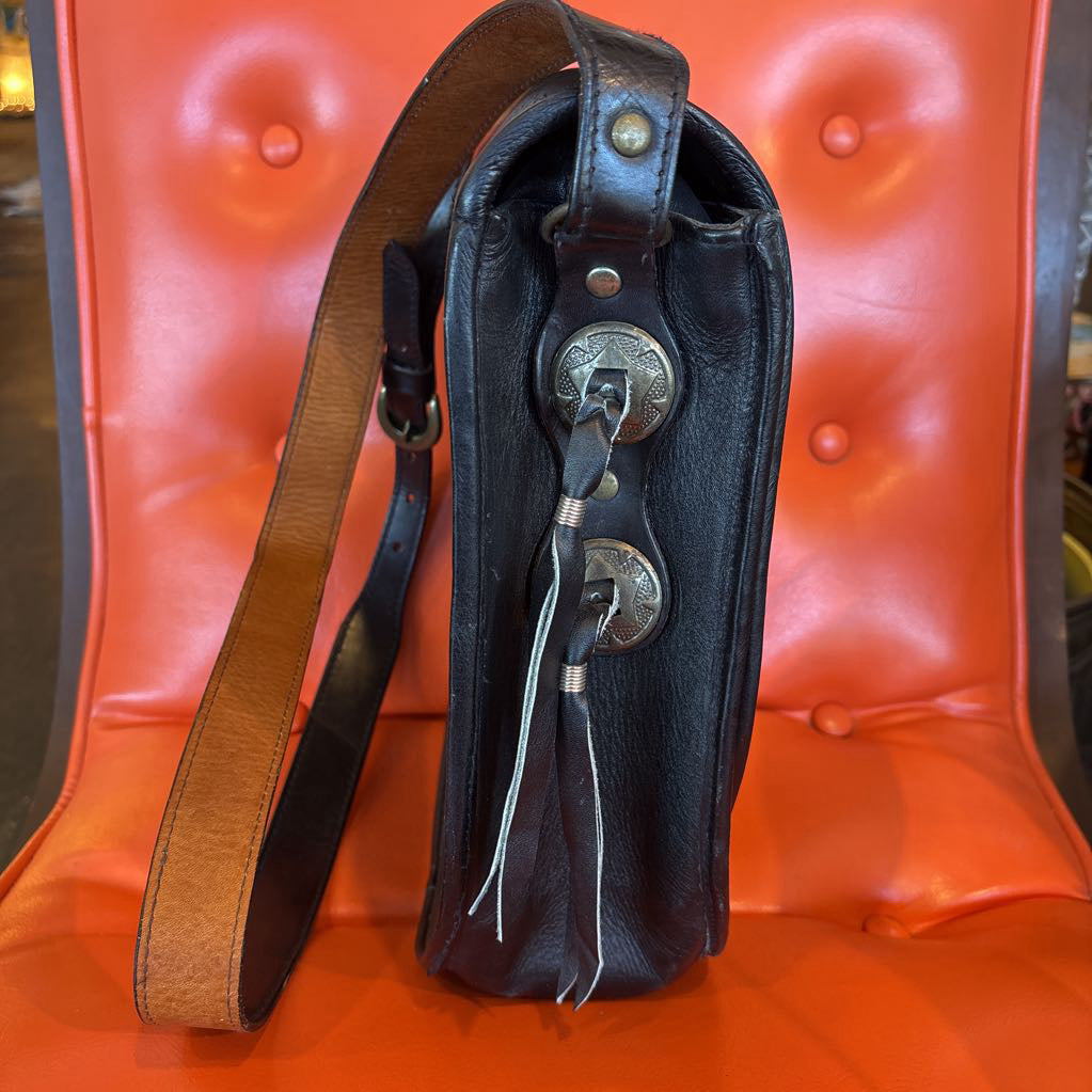 A black leather belt bag with silver hardware and fringe pieces. This bag sits on an orange cushion in a room setting.