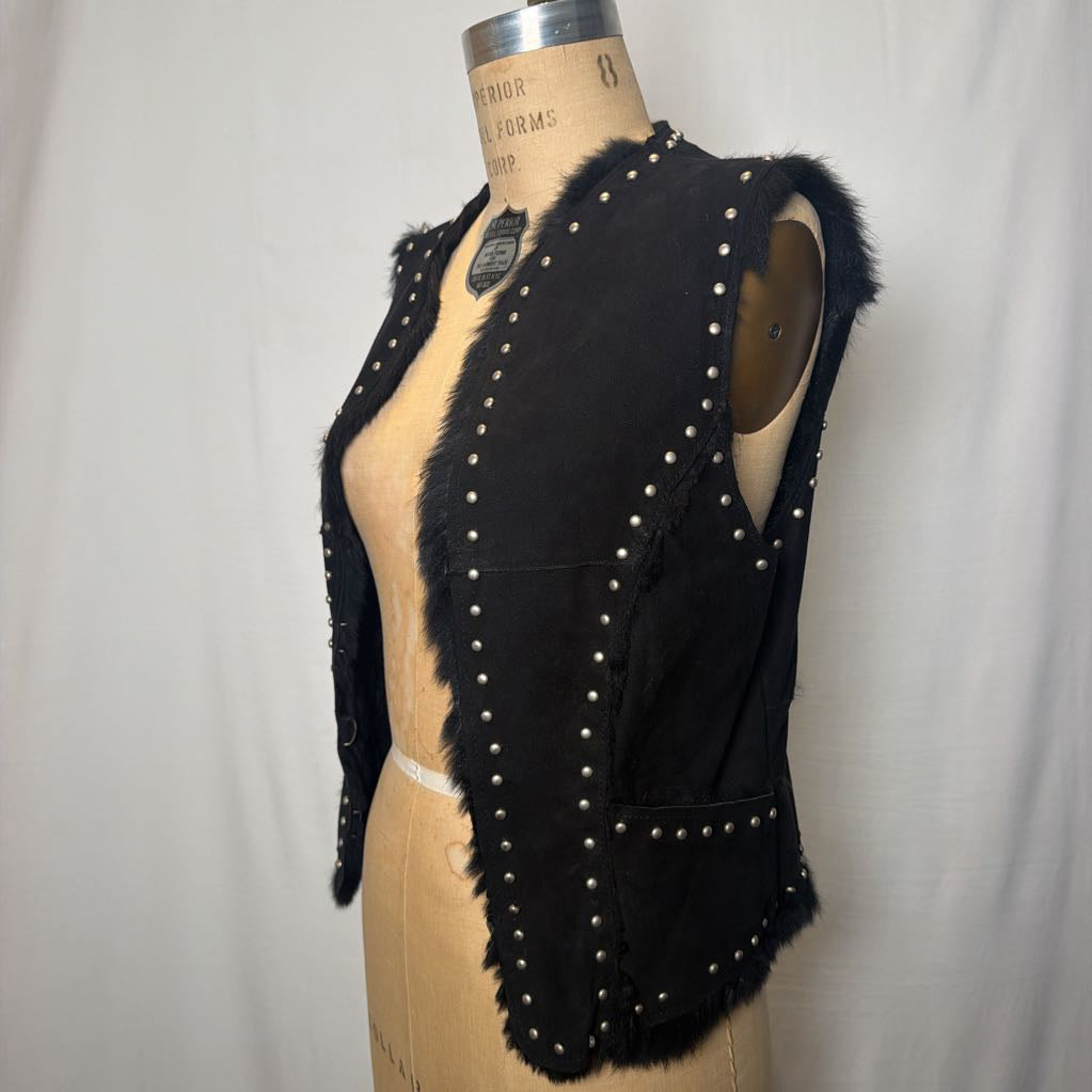 A black suede vest with silver button embellishments and black fur on the inside fluffing out. It's worn on an antique mannequin that sits in front of a white fabric wall. 