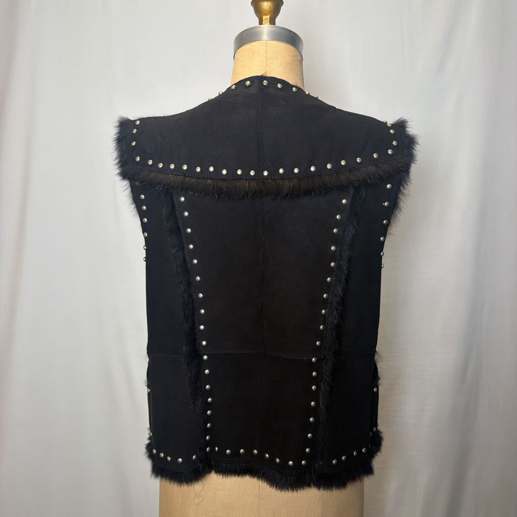 A black suede vest with silver button embellishments and black fur on the inside fluffing out. It's worn on an antique mannequin that sits in front of a white fabric wall. 
