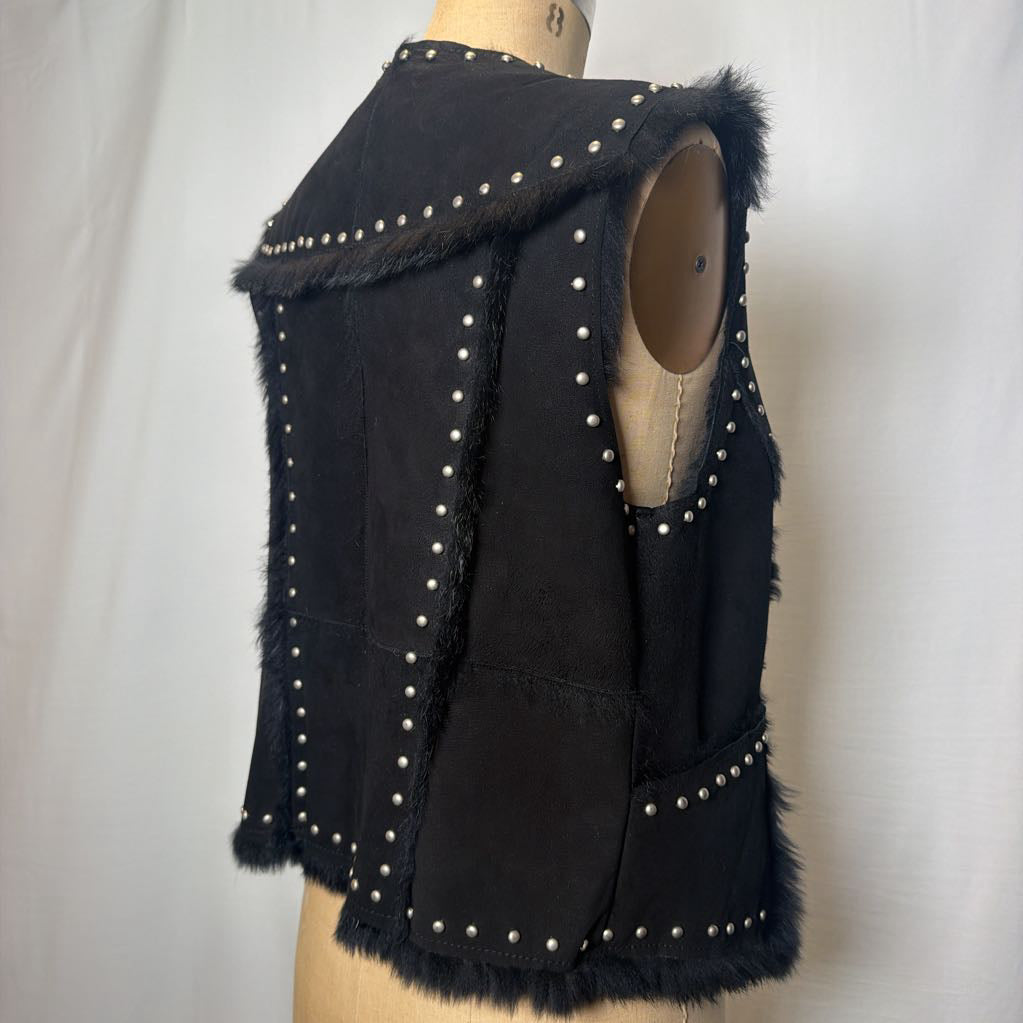 A black suede vest with silver button embellishments and black fur on the inside fluffing out. It's worn on an antique mannequin that sits in front of a white fabric wall. 