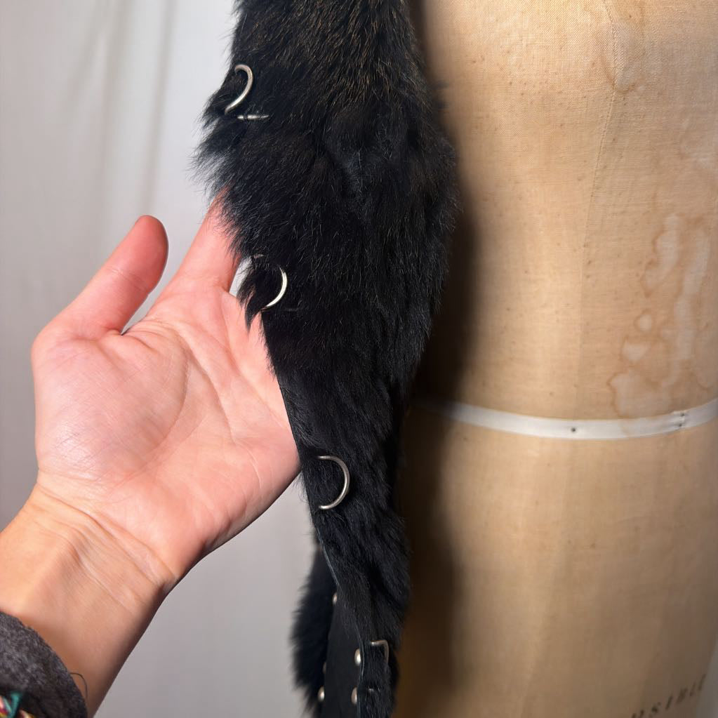 A black suede vest with silver button embellishments and black fur on the inside fluffing out. It's worn on an antique mannequin that sits in front of a white fabric wall. 