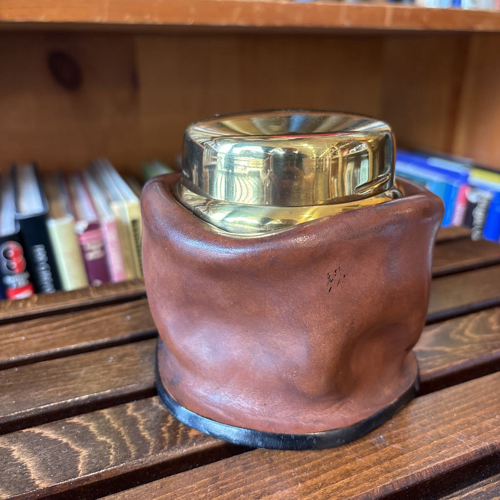 A brass and leather round object on a wooden slat bench in front of books. 