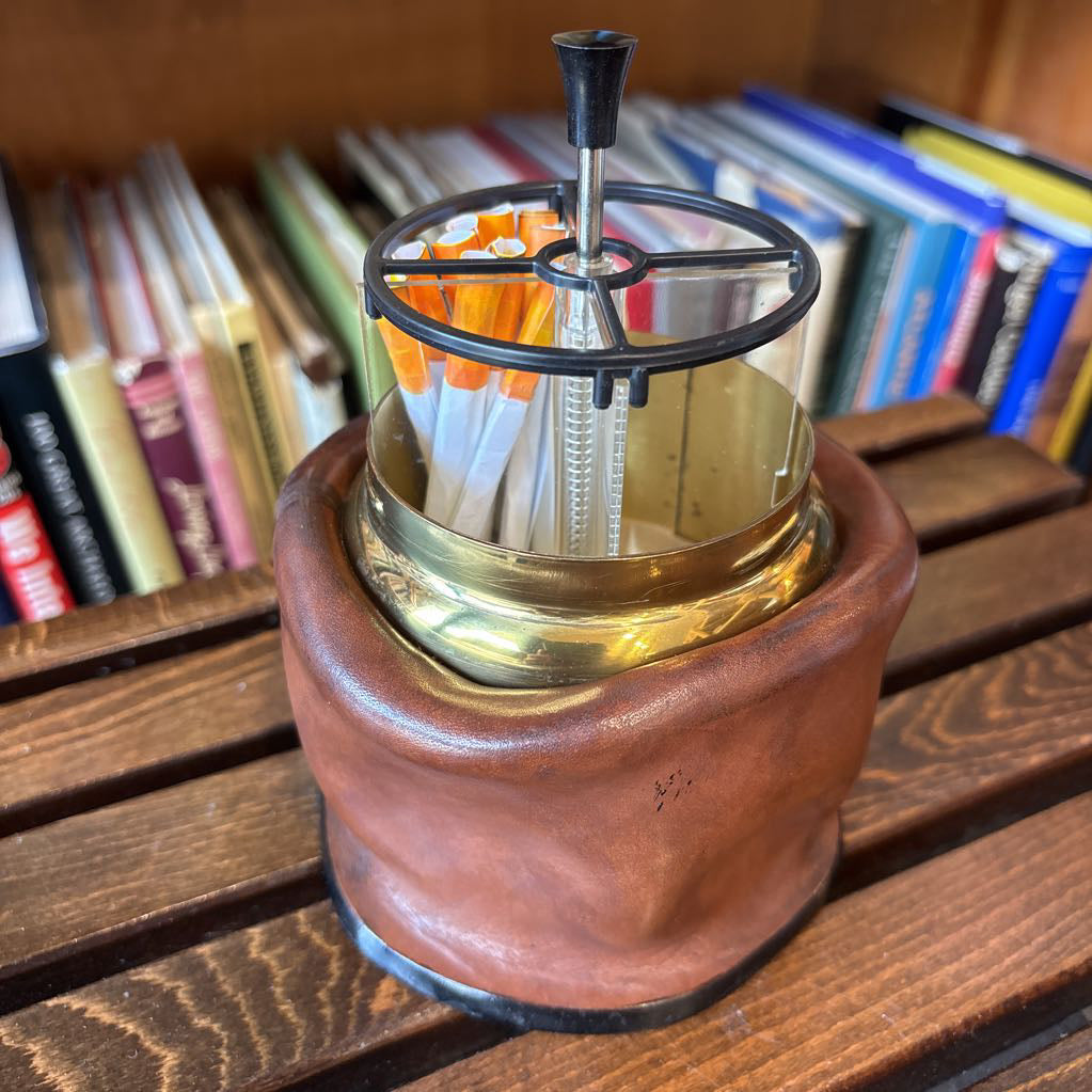 A brass and black metal cigarette holder with orange and white filter tips, set inside a brown leather-like holder on a wooden table with a blurred bookshelf background.