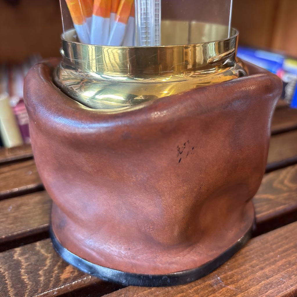 A brass and black metal cigarette holder with orange and white filter tips, set inside a brown leather-like holder on a wooden table with a blurred bookshelf background.