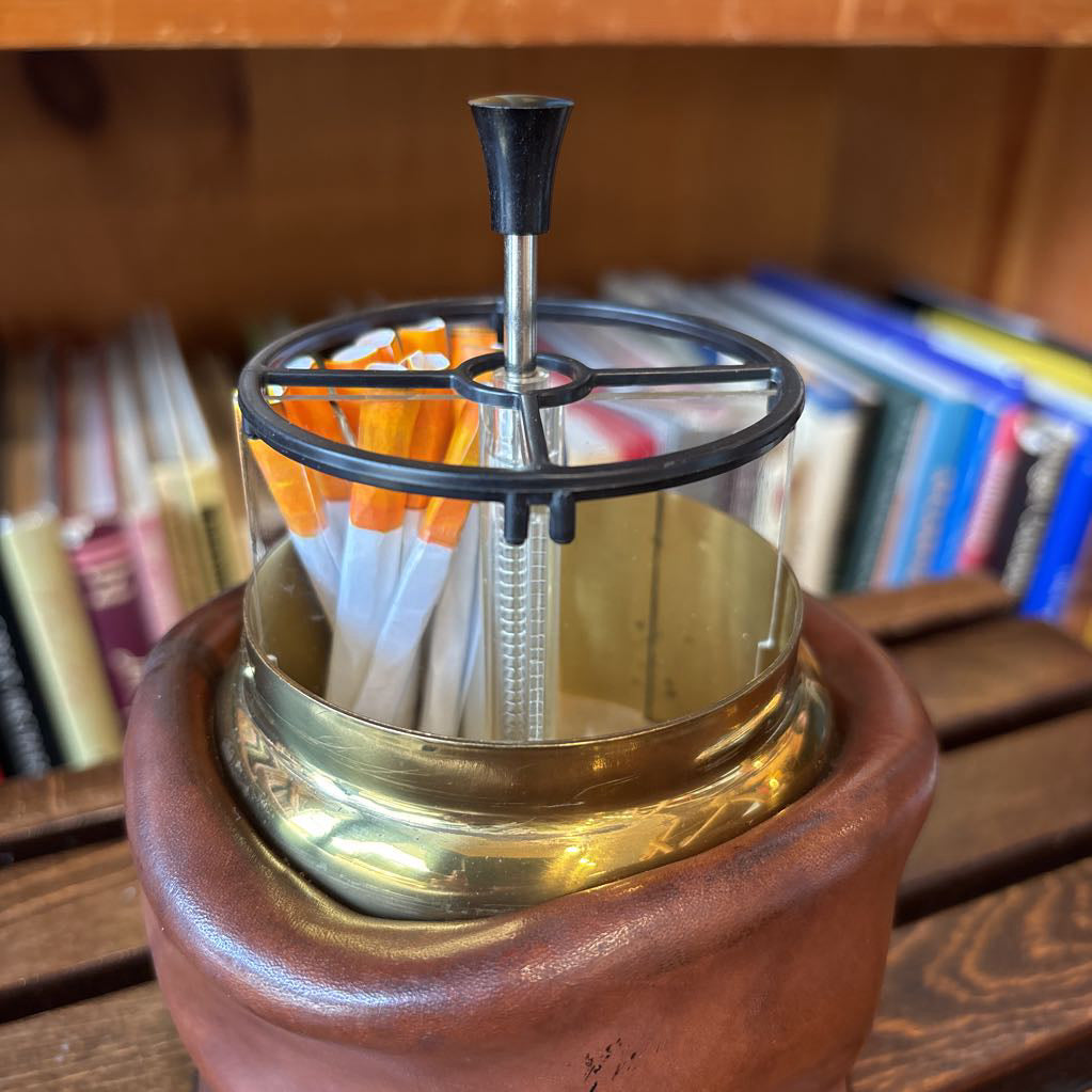 A brass and black metal cigarette holder with orange and white filter tips, set inside a brown leather-like holder on a wooden table with a blurred bookshelf background.