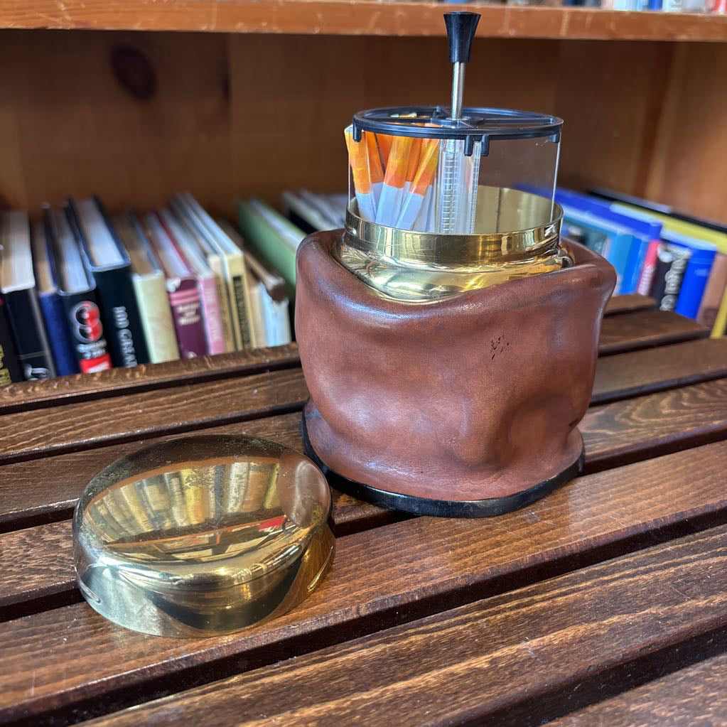 A brass and black metal cigarette holder with orange and white filter tips, set inside a brown leather-like holder on a wooden table with a blurred bookshelf background.