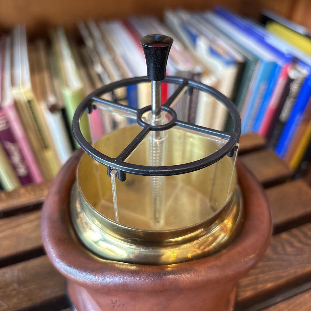 A brass and black metal cigarette holder set inside a brown leather-like holder on a wooden table with a blurred bookshelf background.