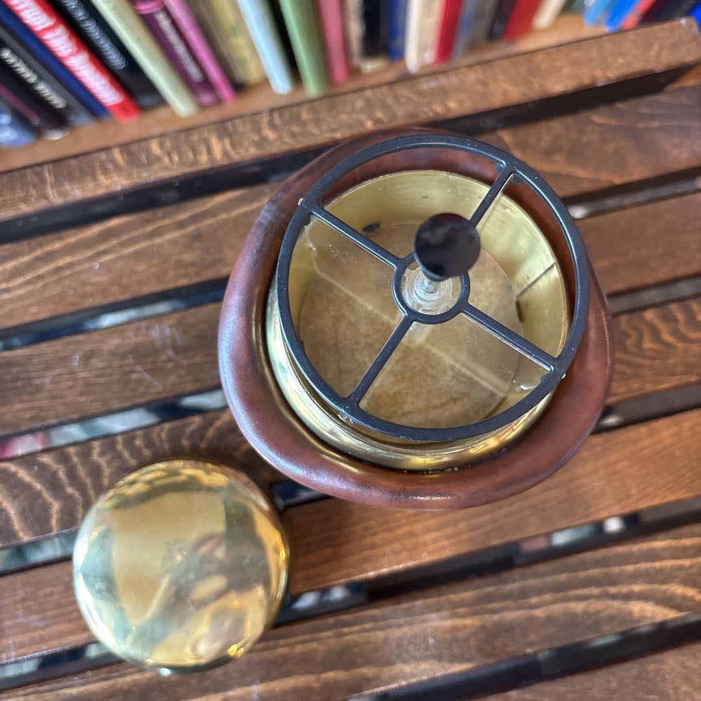 A brass and black metal cigarette holder set inside a brown leather-like holder on a wooden table with a blurred bookshelf background.