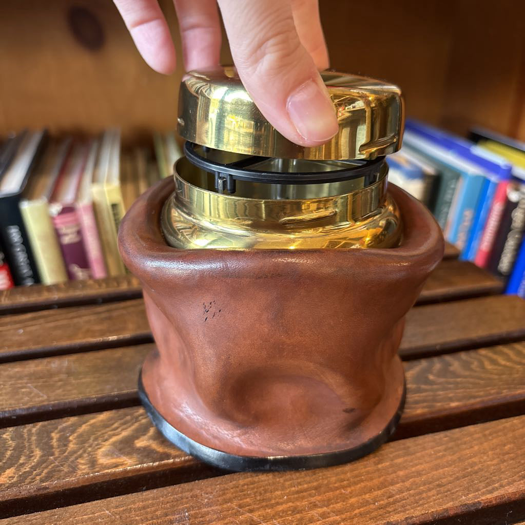 A brass and black metal cigarette holder set inside a brown leather-like holder on a wooden table with a blurred bookshelf background.