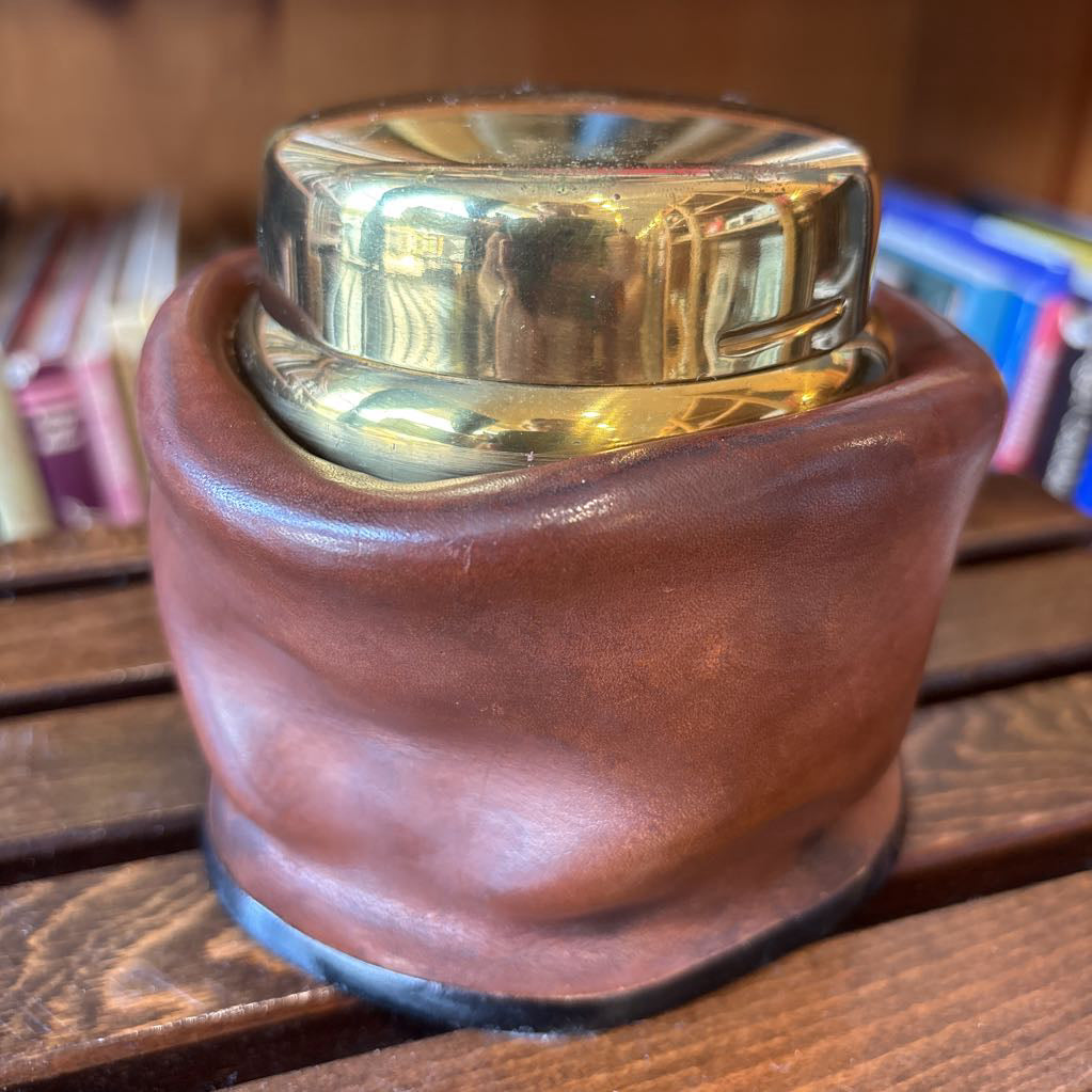 A brass and leather round object on a wooden slat bench in front of books. 