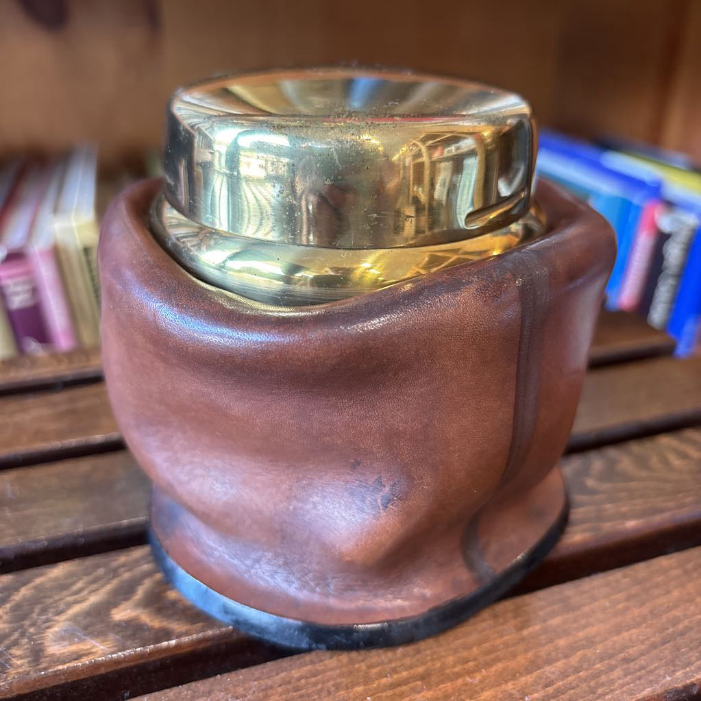 A brass and leather round object on a wooden slat bench in front of books. 