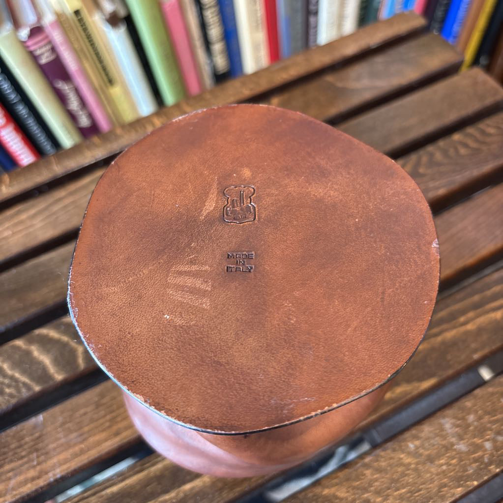 A close-up of the circular brown leather lid of a vintage leather case, featuring an embossed monogram logo and maker's mark, resting on a dark wooden slatted table with a blurred background of books.