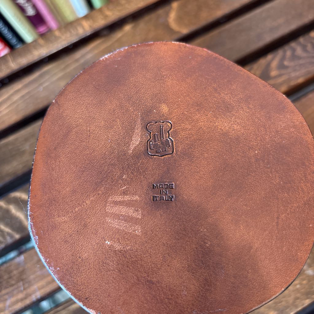 A close-up of the circular brown leather lid of a vintage leather case, featuring an embossed monogram logo and maker's mark, resting on a dark wooden slatted table with a blurred background of books.
