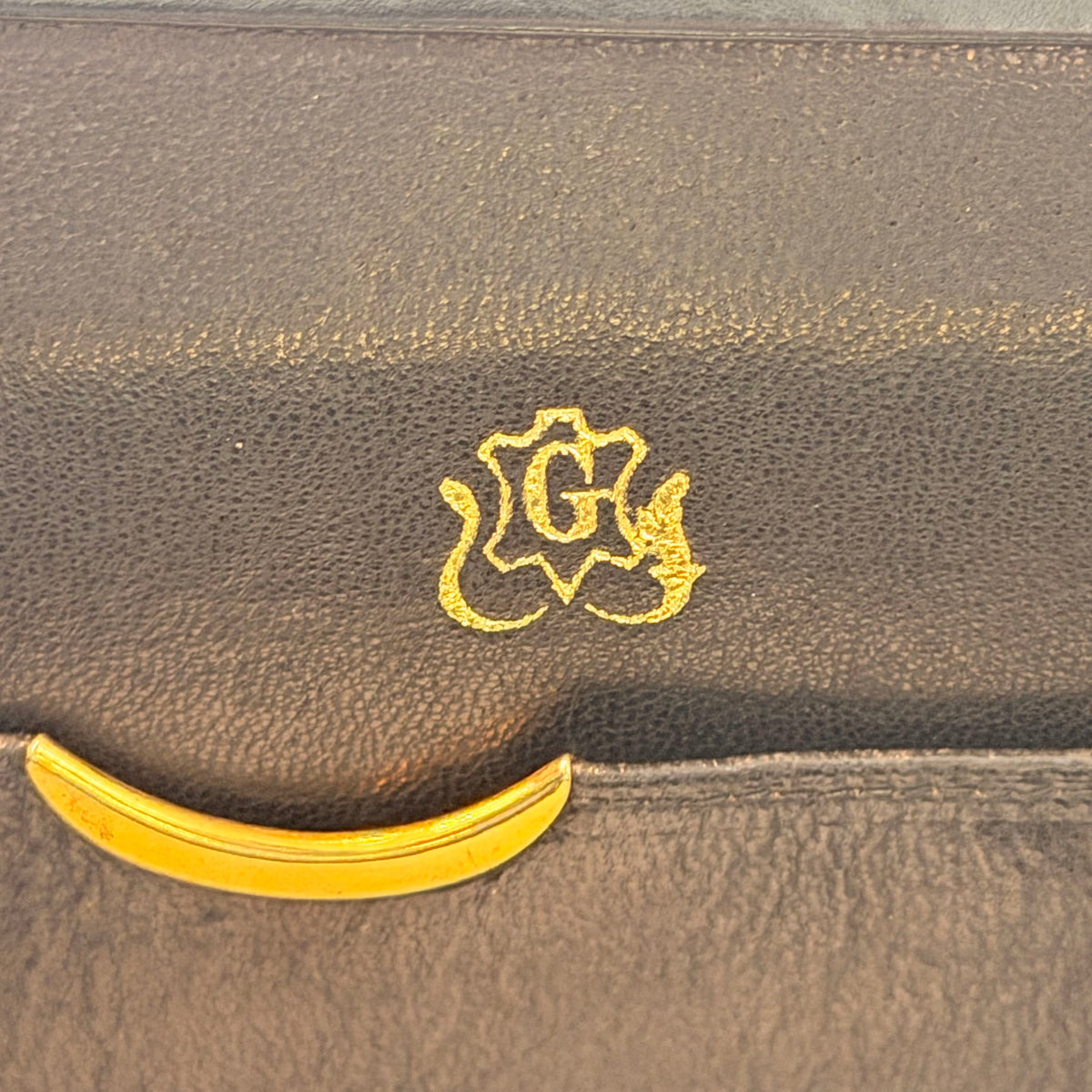 Close-up of a dark brown textured leather item featuring a gold crest logo with the letter G and a gold curved accent at the bottom.