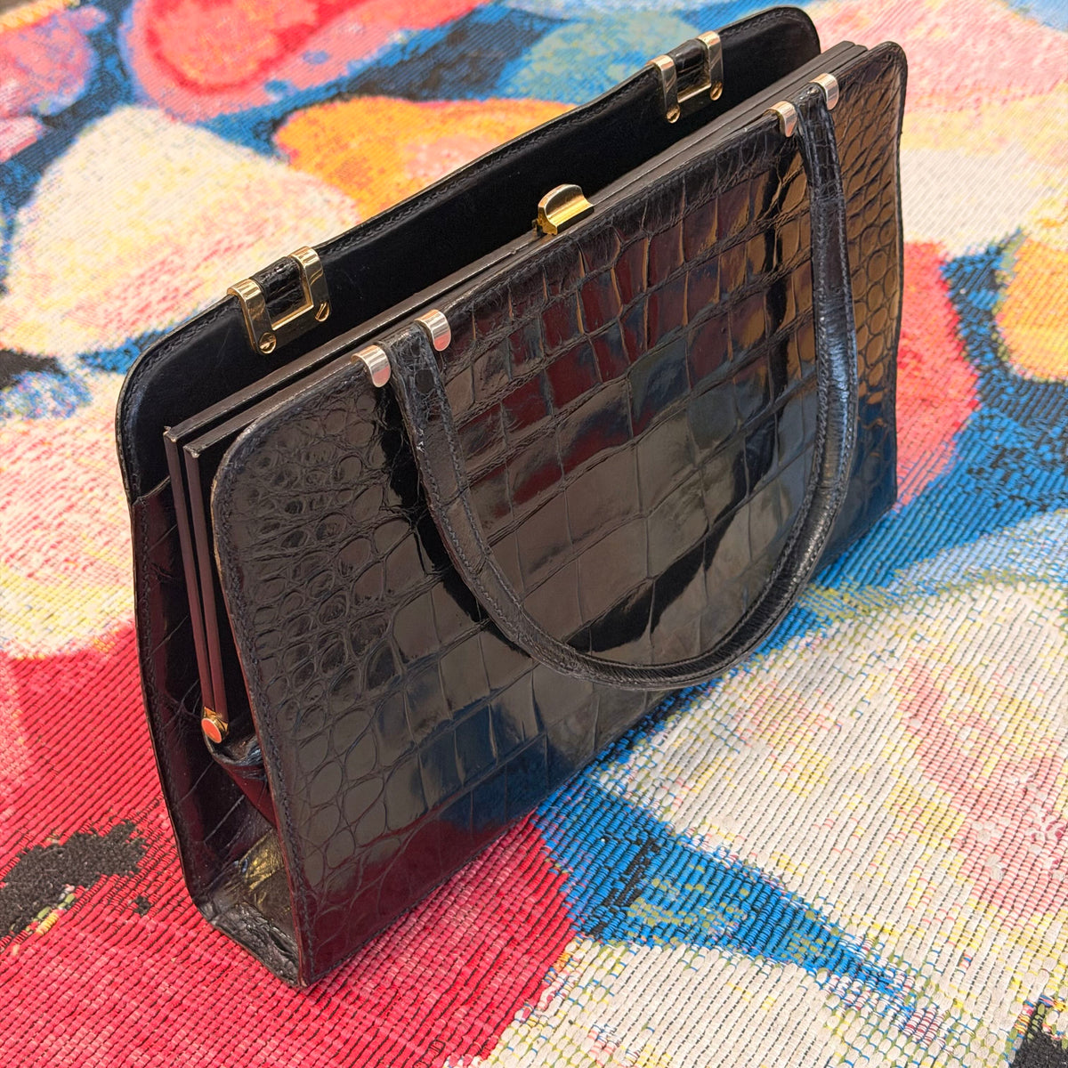 A black crocodile-embossed leather clutch handbag with gold-tone hardware, resting on a colorful woven textile background.