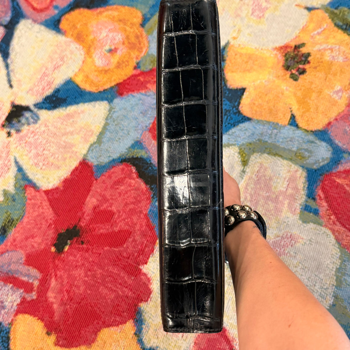 A black crocodile-embossed leather clutch or wallet held vertically by a person's hand, set against a vibrant, multicolored floral patterned fabric background.