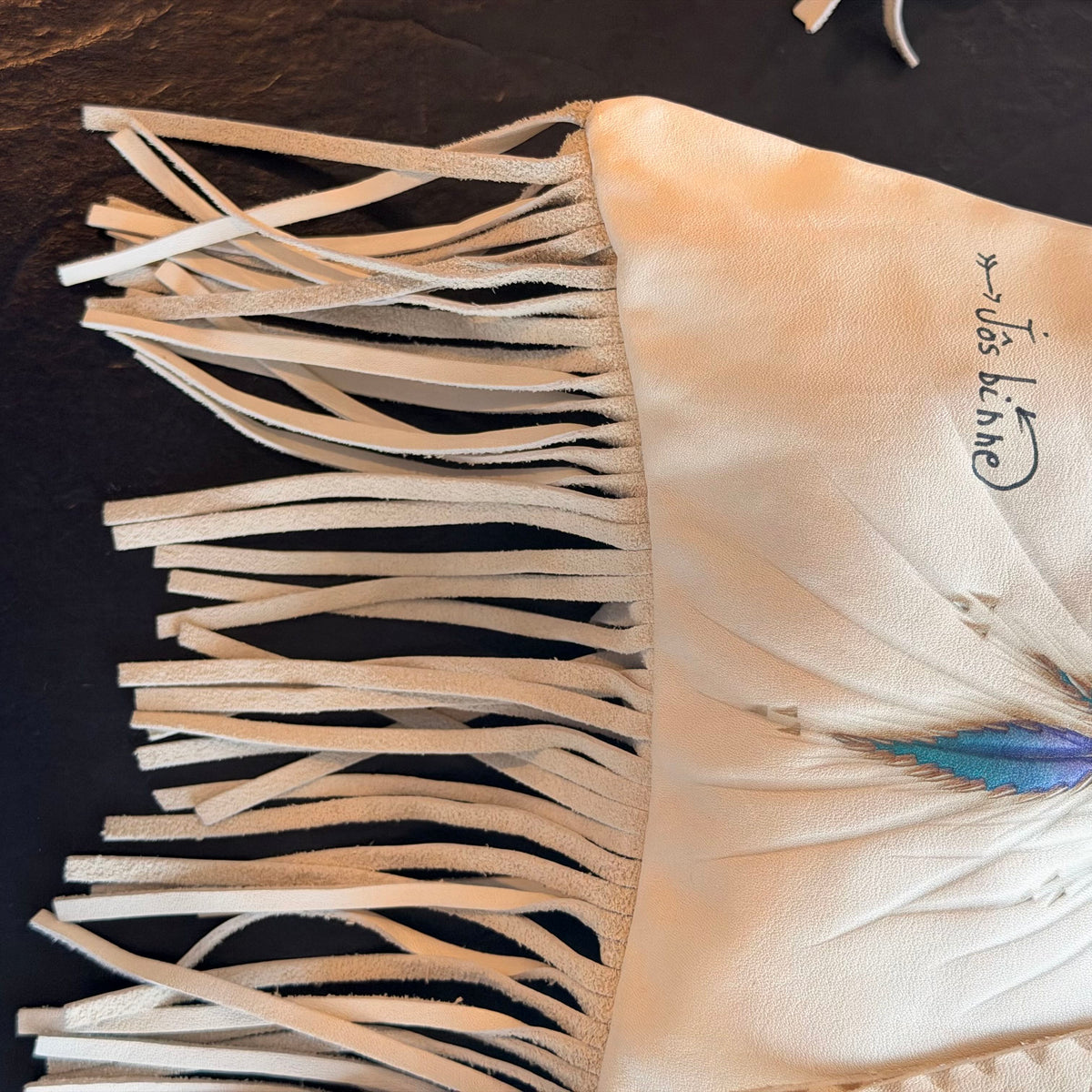 Close-up of a cream-colored leather fringe garment, likely a skirt or apron, featuring a blue and purple feather accent and the handwritten text 'Jésé l’abé' on the right side, laid flat on a dark textured surface.