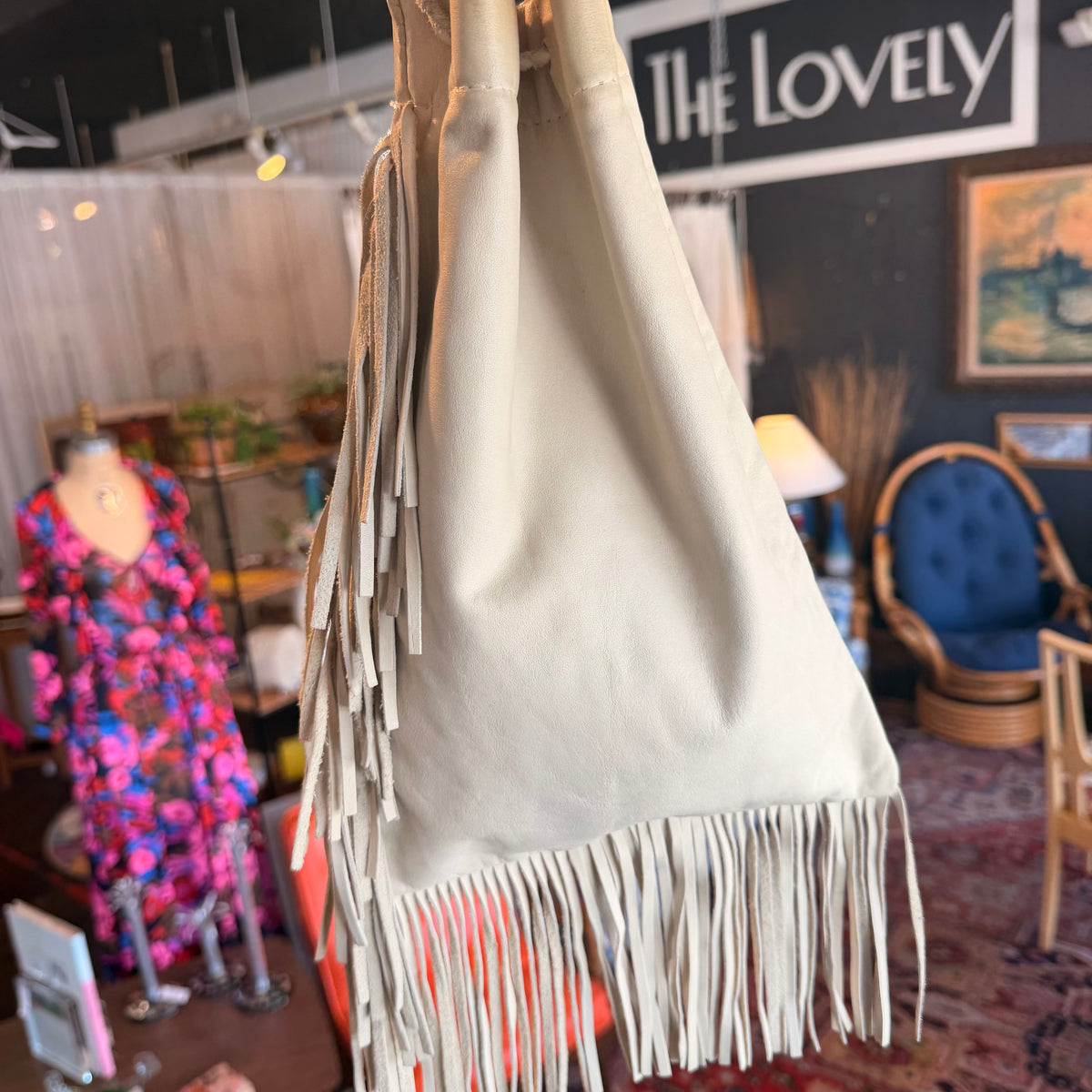 A cream-colored suede fringe bag hanging in a boutique setting, with a mannequin wearing a colorful floral dress visible in the blurred background.