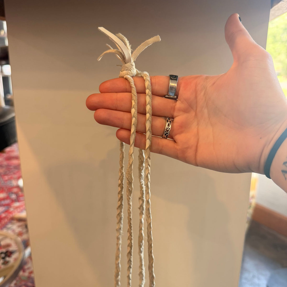 A hand holding a cream-colored braided cord or rope with a knotted top, shown against an indoor background with a beige wall and some room details visible.