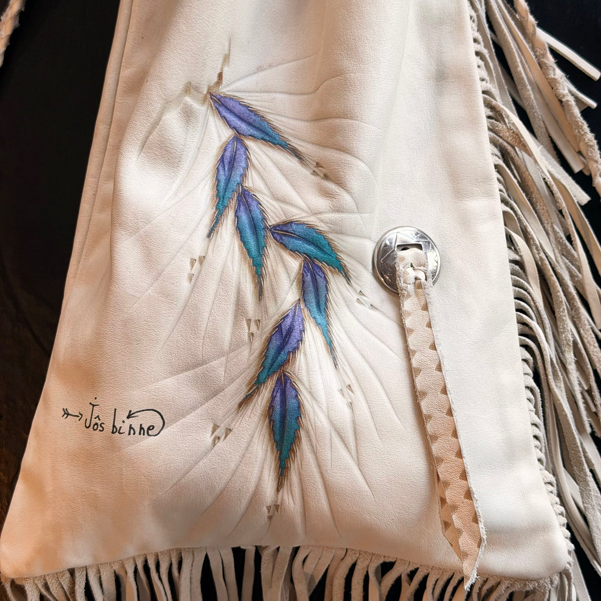 Close-up of a cream-colored leather fringe garment or bag featuring embroidered blue and purple leaf motifs, a silver-tone snap closure, and a small black handwritten signature near the lower left.