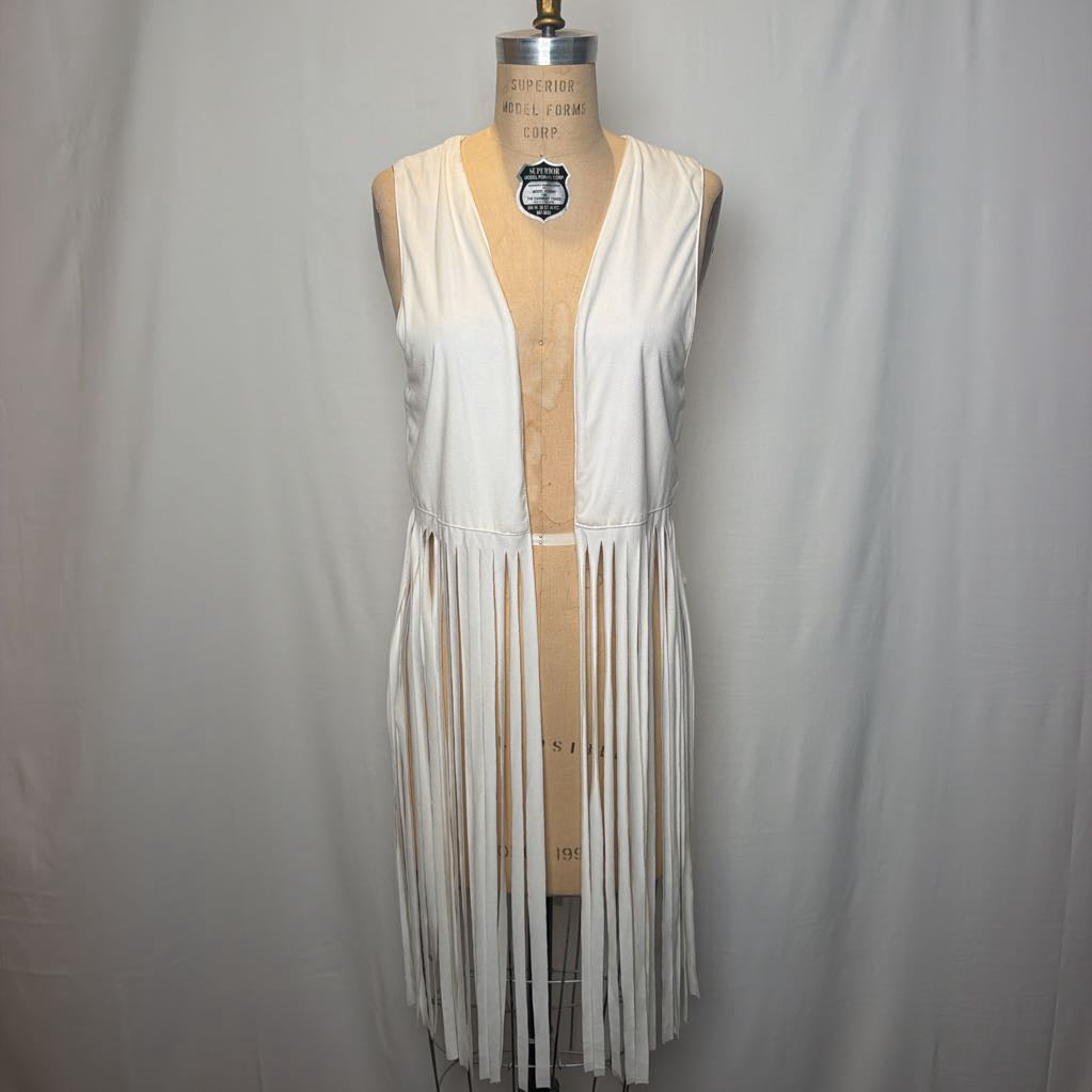 A white vest with long fringe on an antique mannequin in front of a white fabric wall.