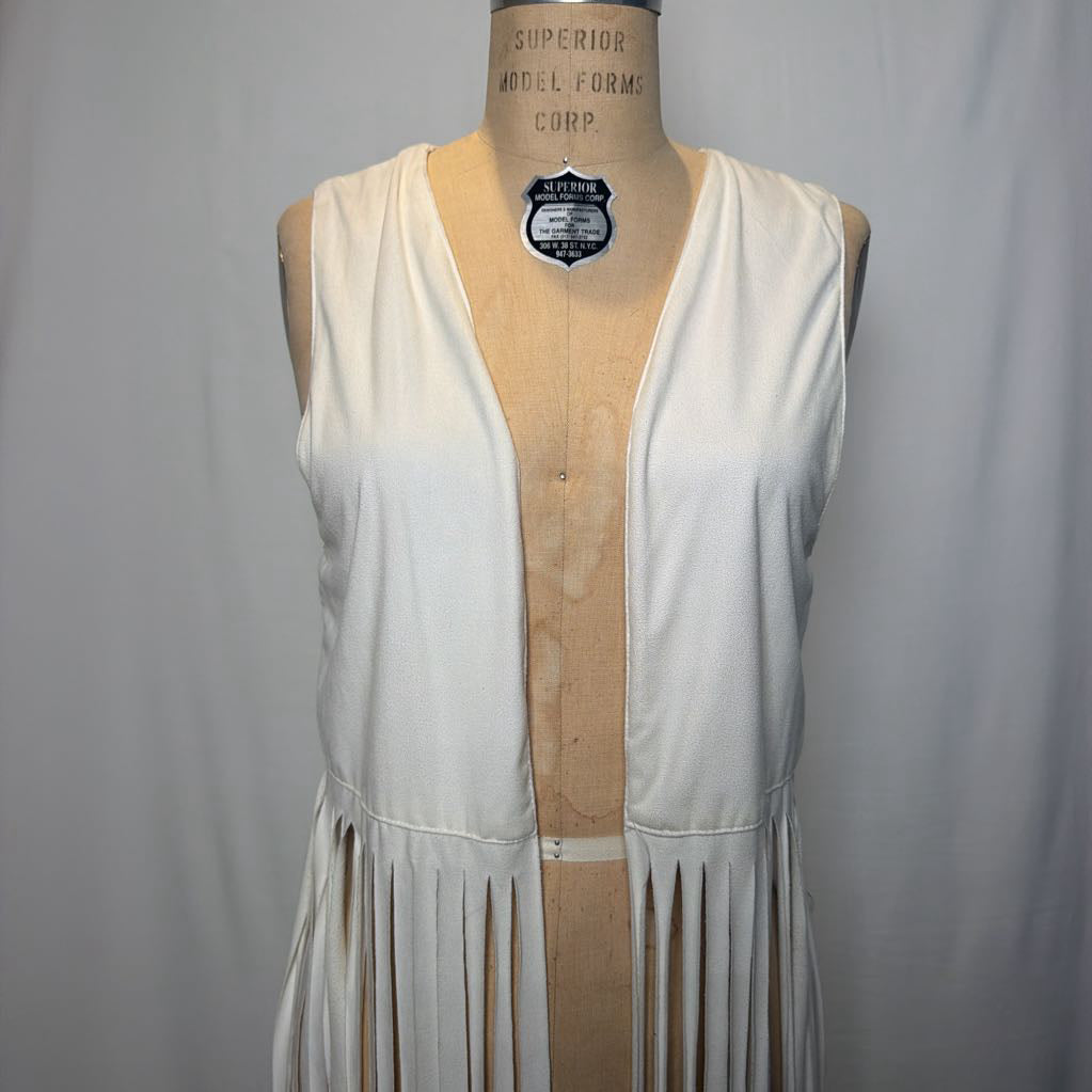 A white vest with long fringe on an antique mannequin in front of a white fabric wall.