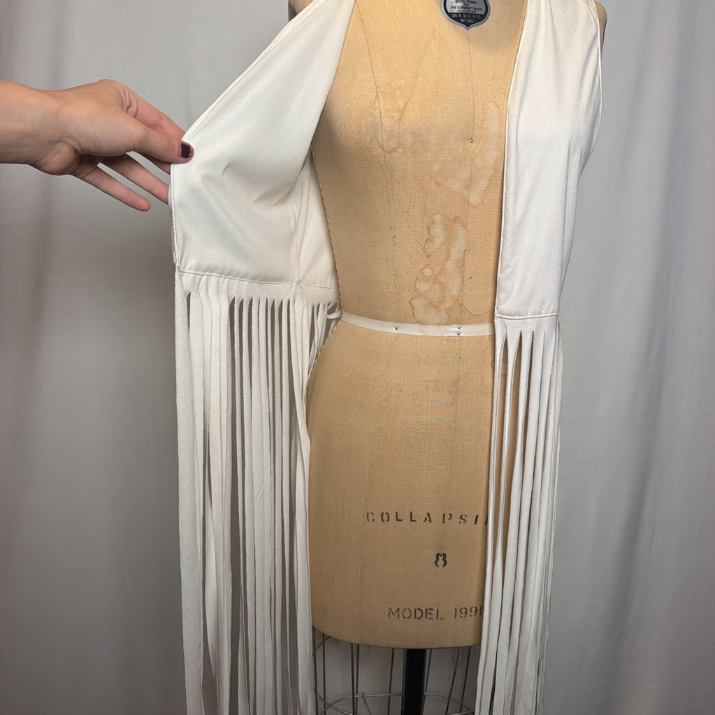 A white vest with long fringe on an antique mannequin in front of a white fabric wall.