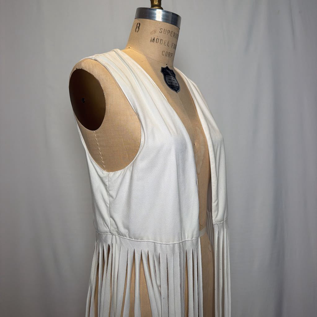 A white vest with long fringe on an antique mannequin in front of a white fabric wall.
