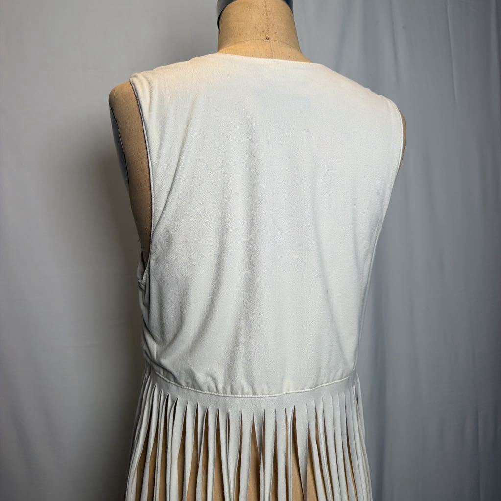 A white vest with long fringe on an antique mannequin in front of a white fabric wall.
