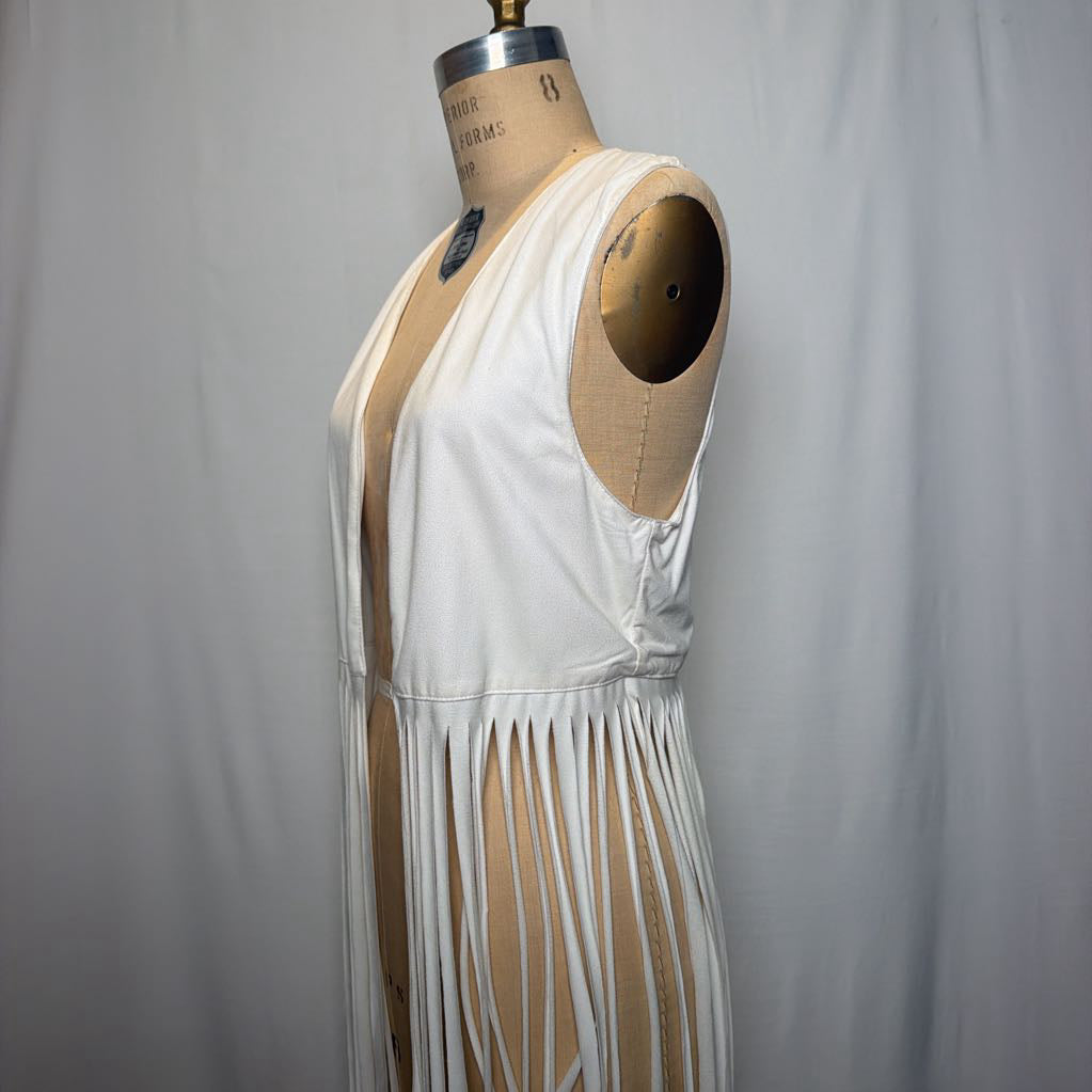 A white vest with long fringe on an antique mannequin in front of a white fabric wall.