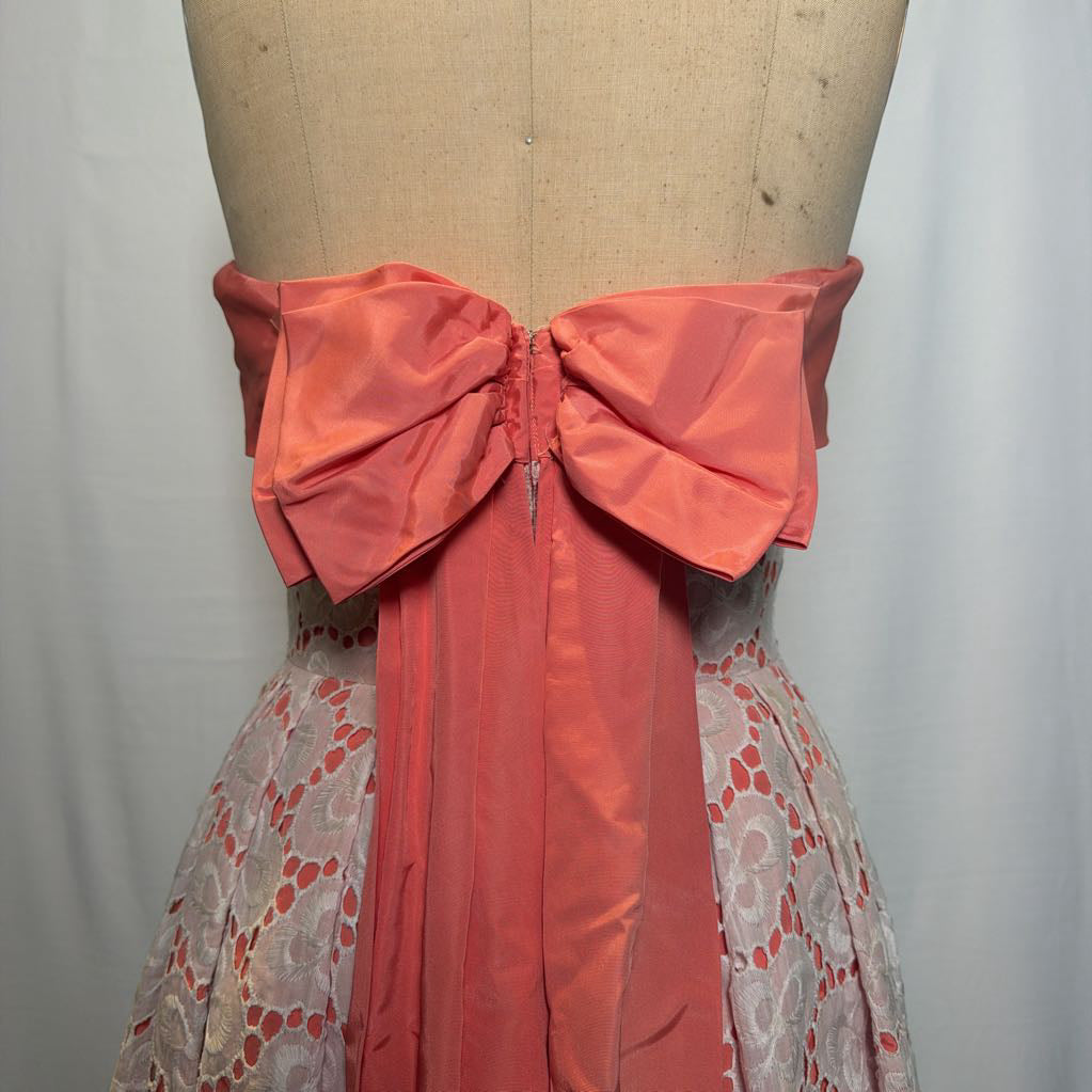 A peach and white lace halter mini dress with a large bow on the back on an antique style mannequin, against a white fabric wall. 