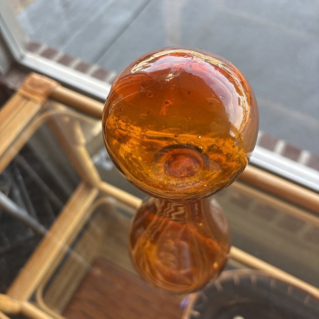 An amber blown glass decanter with a drop shape body and a spherical stopper. Its sitting on a glass table in front of a parking lot. 