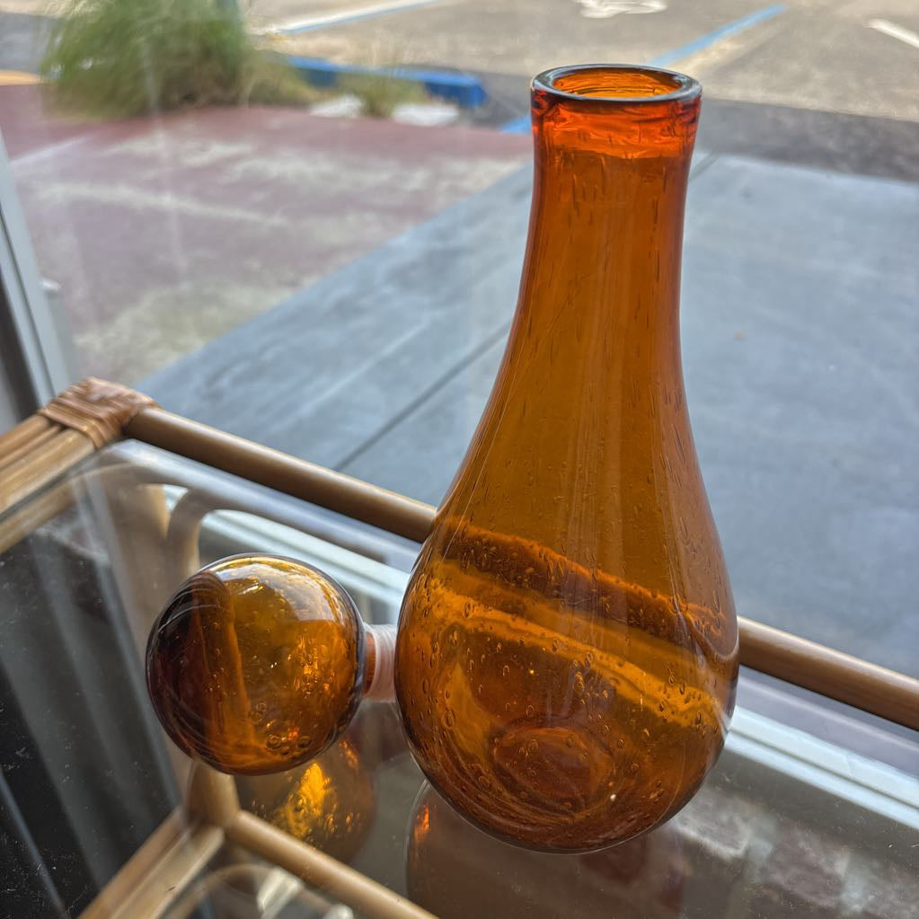 An amber blown glass decanter with a drop shape body and a spherical stopper. Its sitting on a glass table in front of a parking lot. 