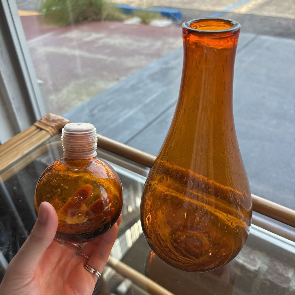 An amber blown glass decanter with a drop shape body and a spherical stopper. Its sitting on a glass table in front of a parking lot. 