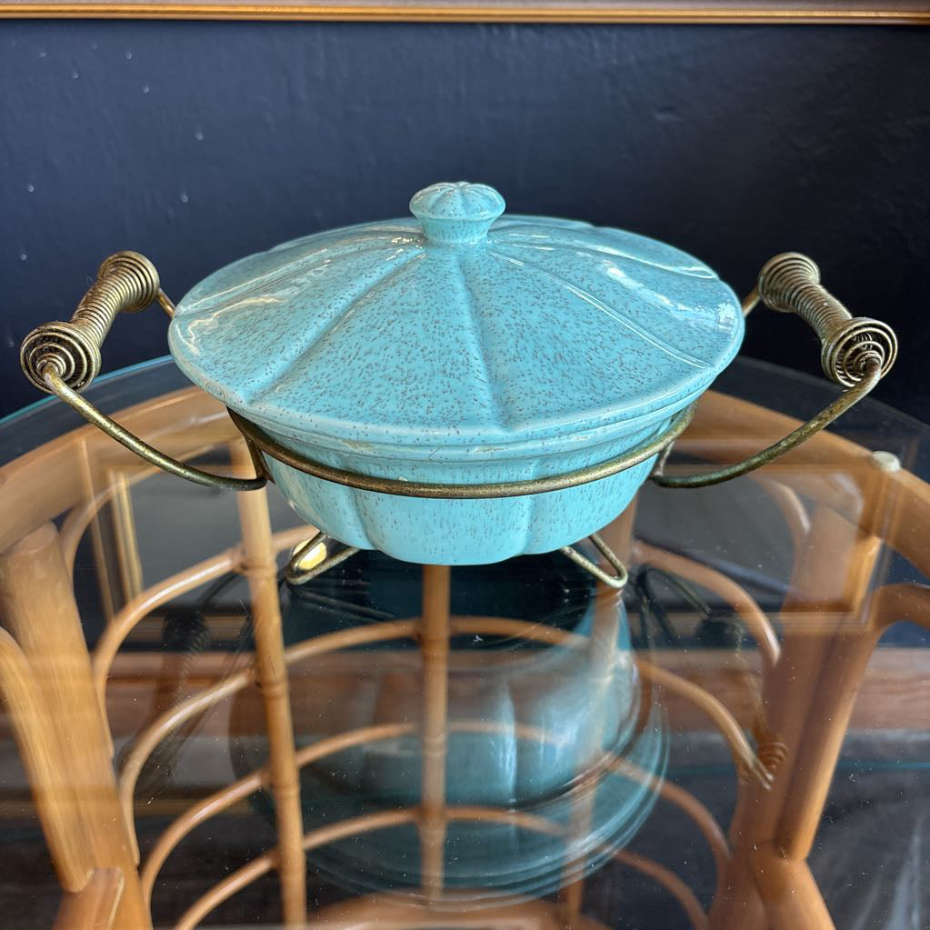 A ceramic turquoise serving bowl and lid in a brass wire caddy with handles on a glass table in front of a black wall.