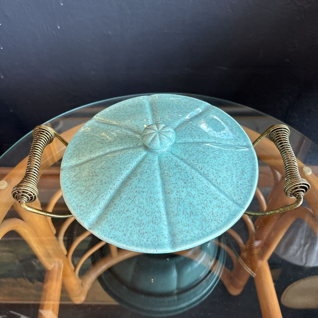 A ceramic turquoise serving bowl and lid in a brass wire caddy with handles on a glass table in front of a black wall.