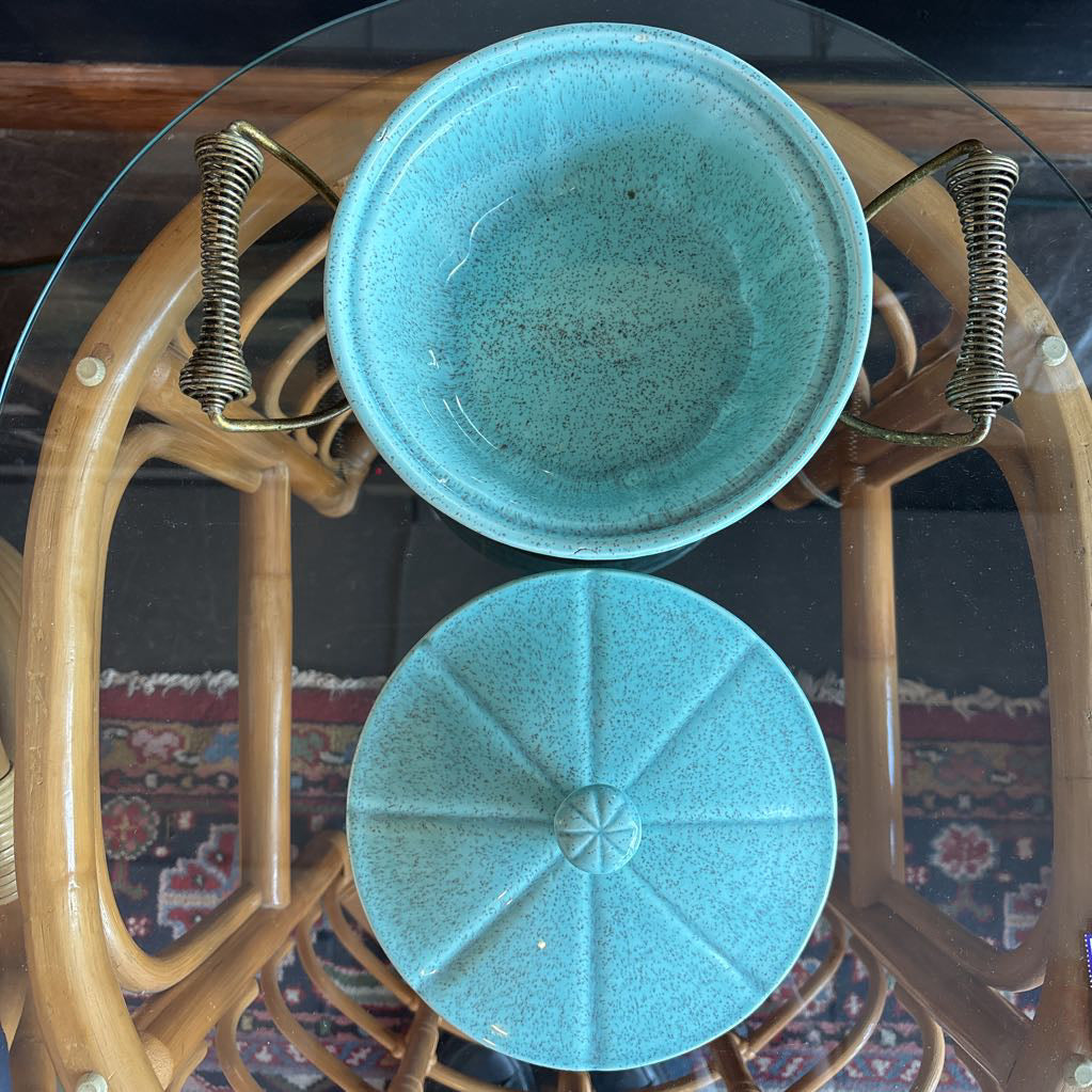 A ceramic turquoise serving bowl and lid in a brass wire caddy with handles on a glass table in front of a black wall.
