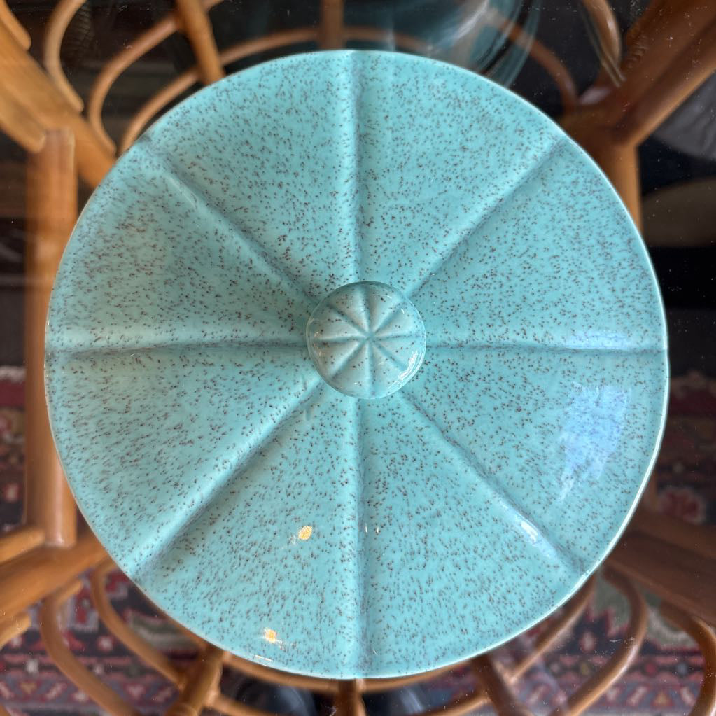 A ceramic turquoise serving bowl and lid in a brass wire caddy with handles on a glass table in front of a black wall.