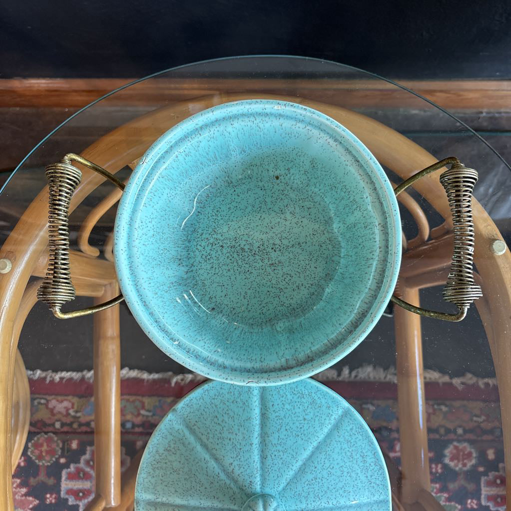 A ceramic turquoise serving bowl and lid in a brass wire caddy with handles on a glass table in front of a black wall.
