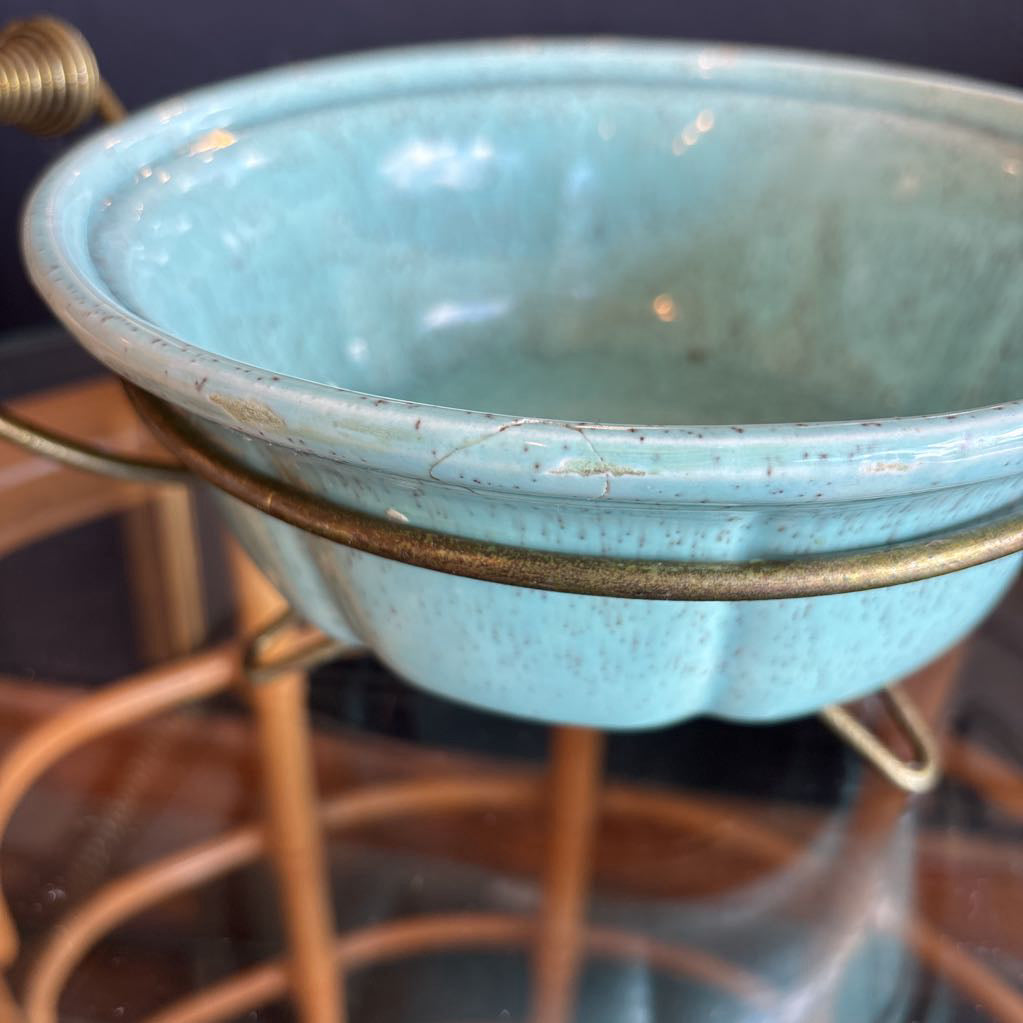 A ceramic turquoise serving bowl and lid in a brass wire caddy with handles on a glass table in front of a black wall.